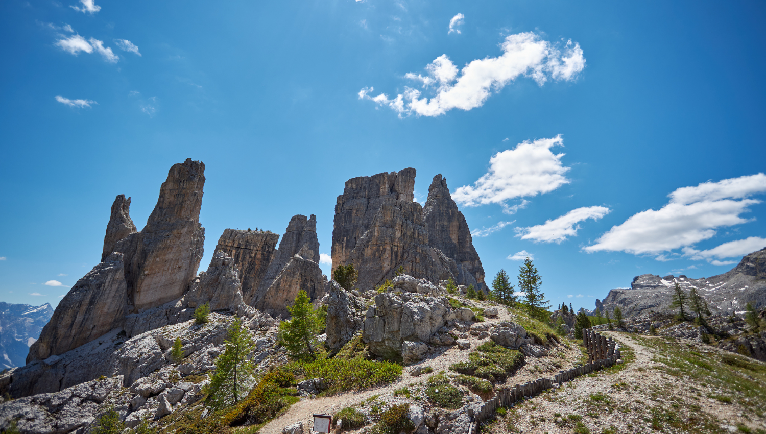 Le Cinque Torri nelle Dolomiti in una giornata soleggiata, con pinnacoli rocciosi maestosi che si ergono contro un cielo blu costellato di nuvole bianche, circondati da pini e un sentiero escursionistico alpino.